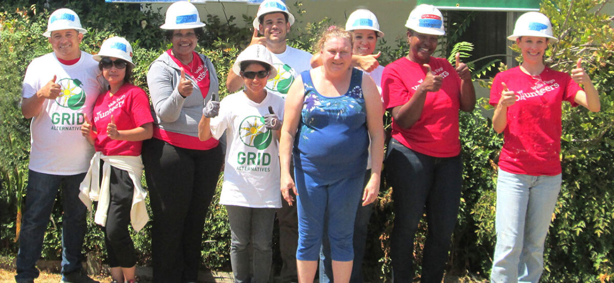 Group of volunteers wearing hard hats and red shirts at a solar energy community event, smiling and giving thumbs up gestures
