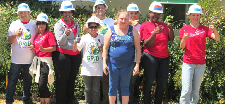 Group of volunteers wearing hard hats and red shirts at a solar energy community event, smiling and giving thumbs up gestures