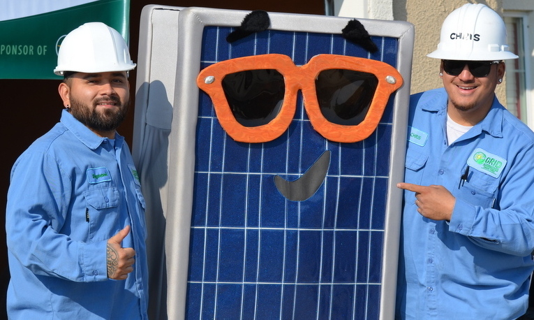 Two workers in hard hats pose with a solar panel decorated with large orange sunglasses and a smile