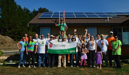 Large group of diverse people posing in front of a house with solar panels, holding a "Bridgeland" banner