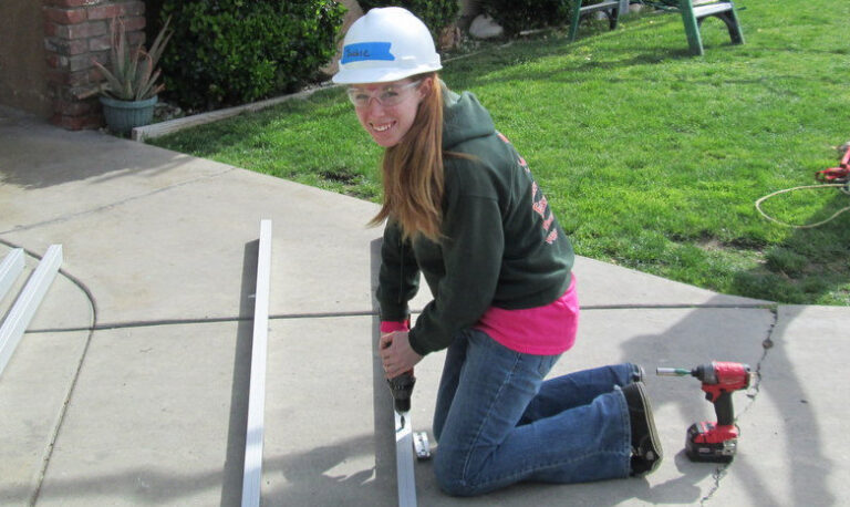 Young woman in hard hat installing solar panel on residential roof with power drill