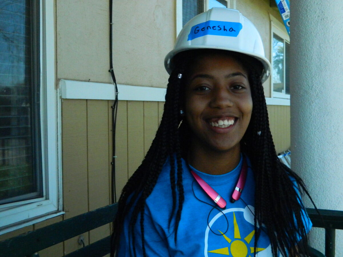Person wearing hard hat labeled "Genesha" and blue shirt with sun logo, smiling at camera