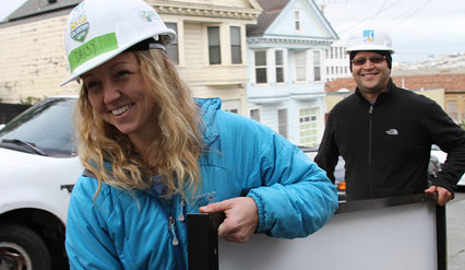 Two people in hard hats and work jackets smile while standing in front of a house under construction