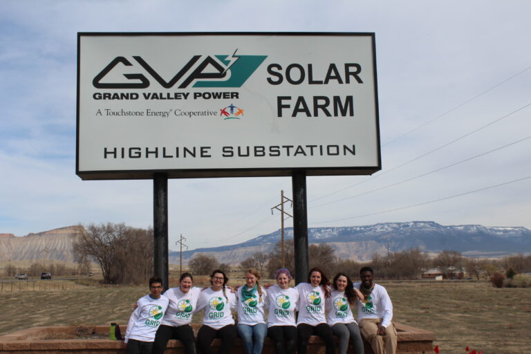 Nine people in GRID t-shirts stand in front of Grand Valley Power Solar Farm Highline Substation sign with mountains in background