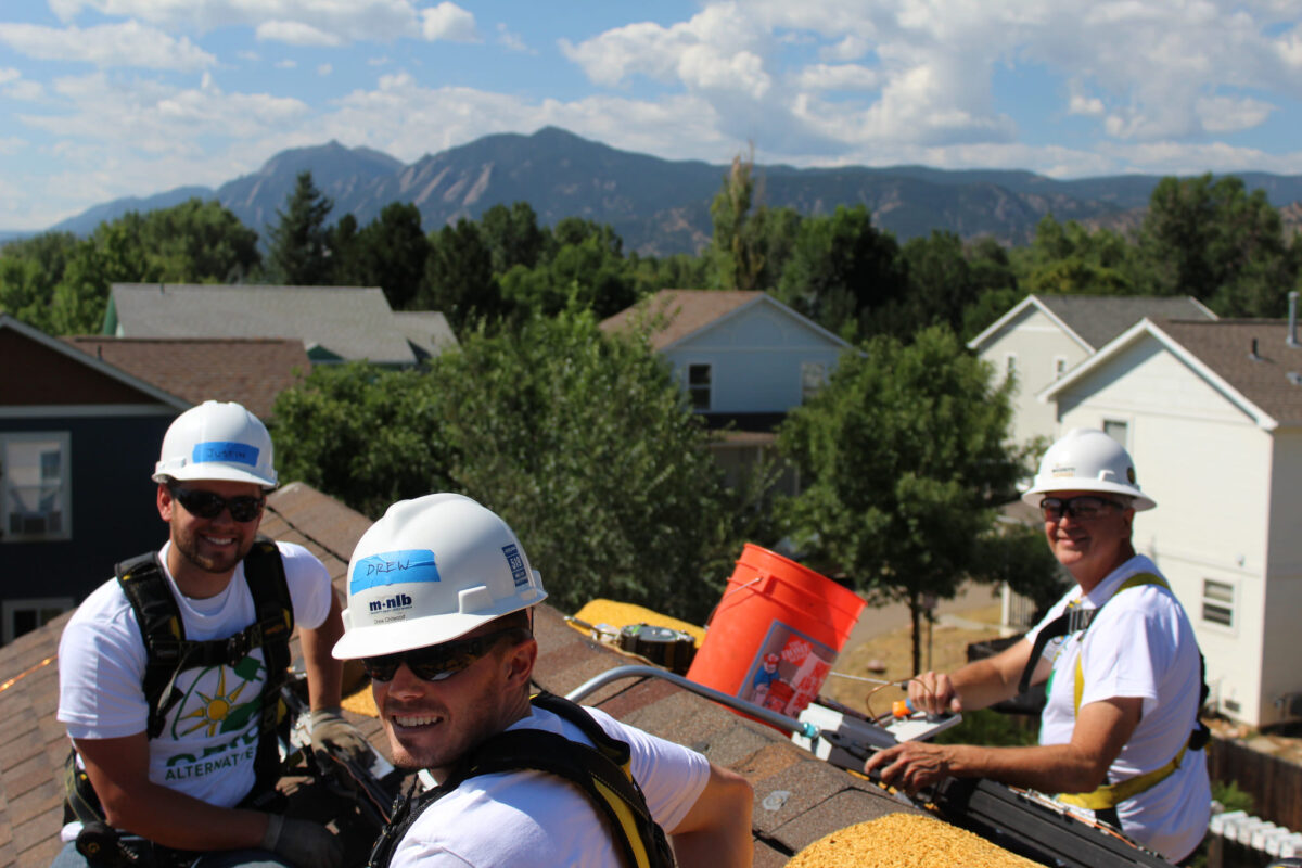 Four volunteers in hard hats and safety gear work on a residential roof with mountains in background