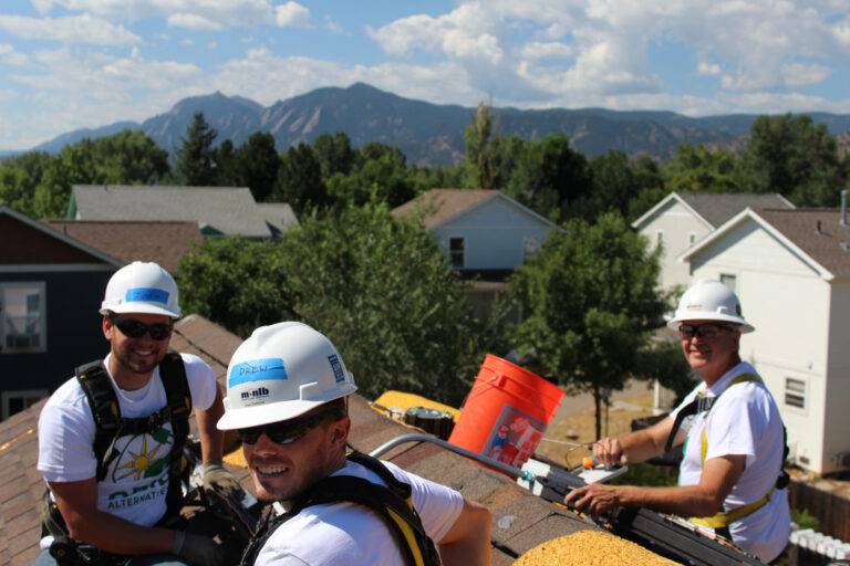 Four volunteers in hard hats and safety gear work on a residential roof with mountains in background