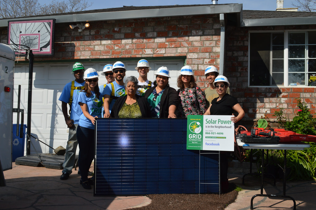 Group of people wearing hard hats holding solar panel with Grid Alternatives sign at residential installation