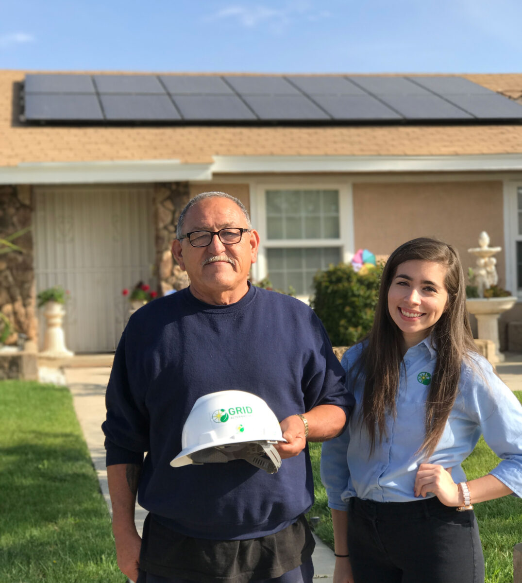 Man and woman holding GRID hard hat in front of house with solar panels on roof