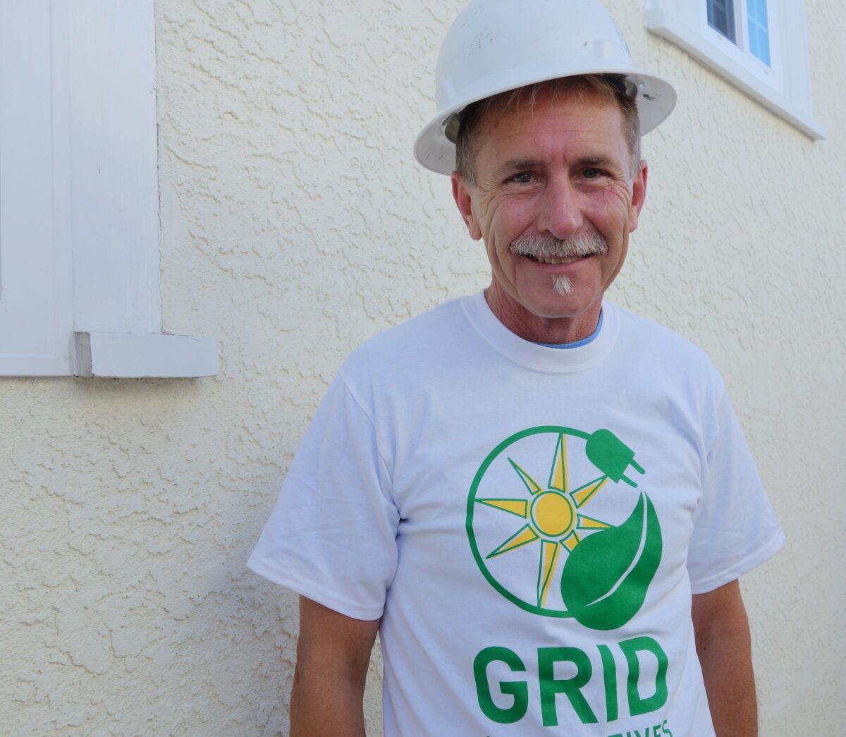 Man in white hard hat wearing GRID sustainability t-shirt smiling at camera against textured white wall