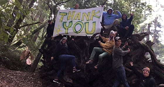Group of people sitting on a large fallen tree holding a "Thank You" sign in a forest