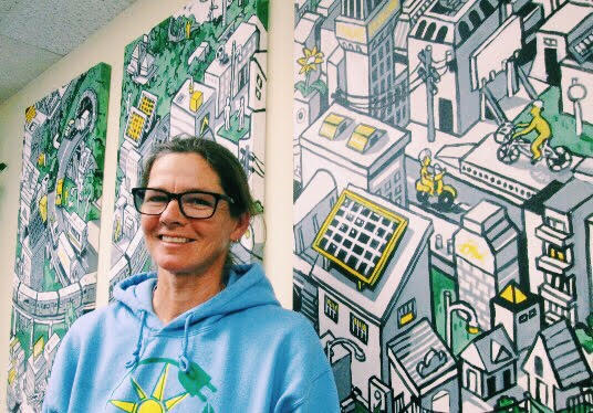 Person wearing blue hoodie smiling in front of colorful illustrated urban murals with buildings and yellow accents