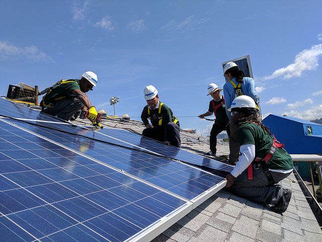 Workers in safety gear installing blue solar panels on a rooftop under clear sky