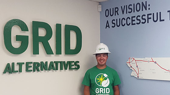 Person in GRID Alternatives shirt and hard hat standing before office wall signage