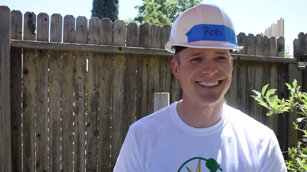 Smiling man wearing hard hat labeled "Rob" and white t-shirt in front of wooden fence
