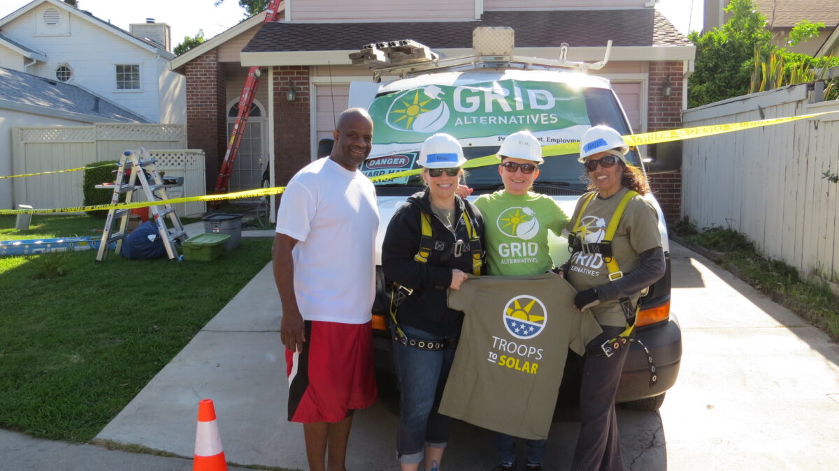 Four people holding "Troops to Solar" sign in front of GRID Alternatives van at residential installation site