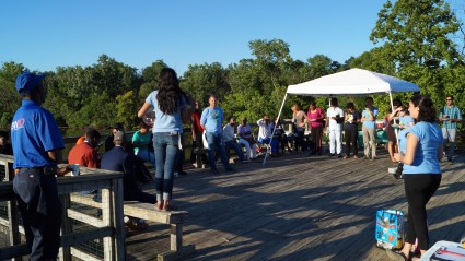 Outdoor community event with attendees gathered under white tent on sunny day, trees in background