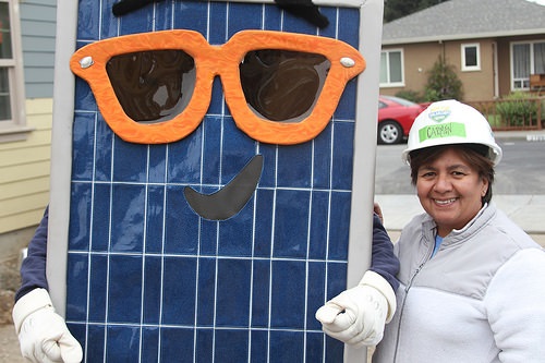 Woman in safety helmet holding large solar panel with oversized orange sunglasses face drawn on it