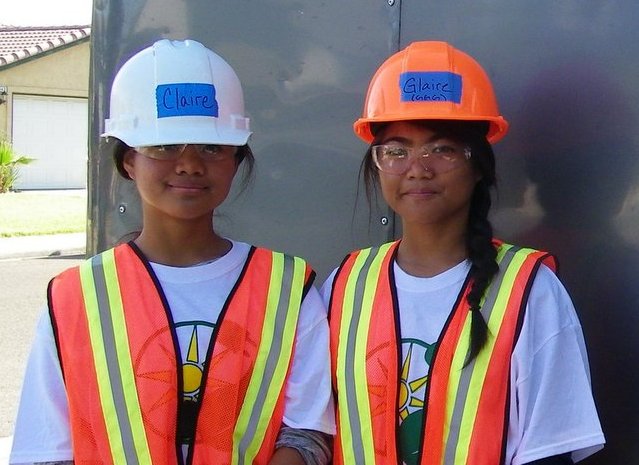 Two young women in safety gear: white and orange hard hats, reflective vests, and glasses, standing together outdoors