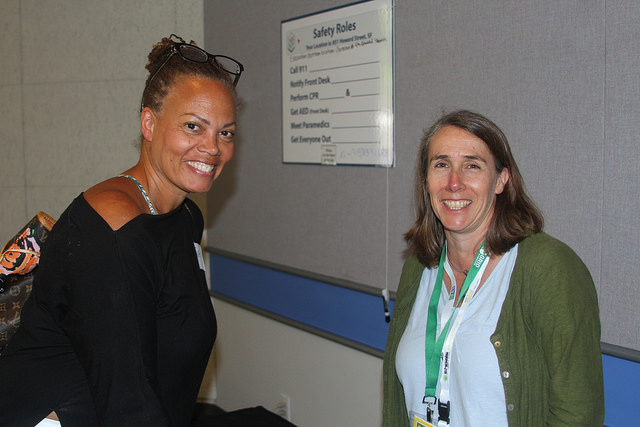 Two smiling women stand together in an indoor setting, one wearing black and the other wearing olive green with a lanyard badge
