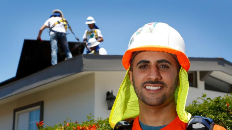 Construction worker in safety gear smiling at camera with team working on roof behind him