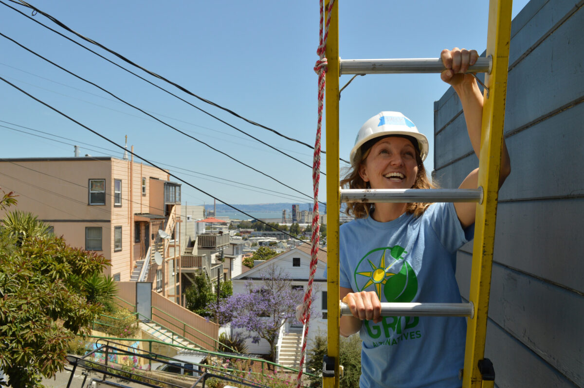 Woman in hard hat climbing yellow ladder on construction site, wearing green solar logo shirt