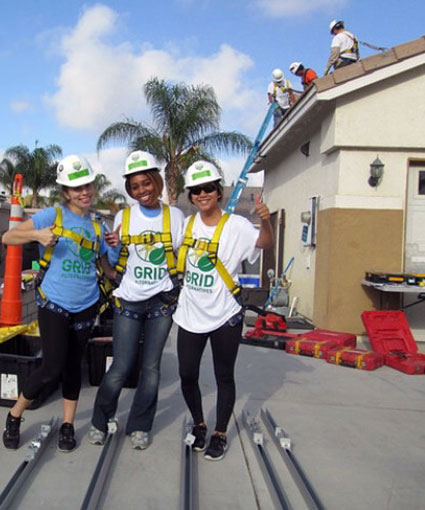 Three volunteers in hard hats and yellow GRID vests on a residential roof during a solar installation project