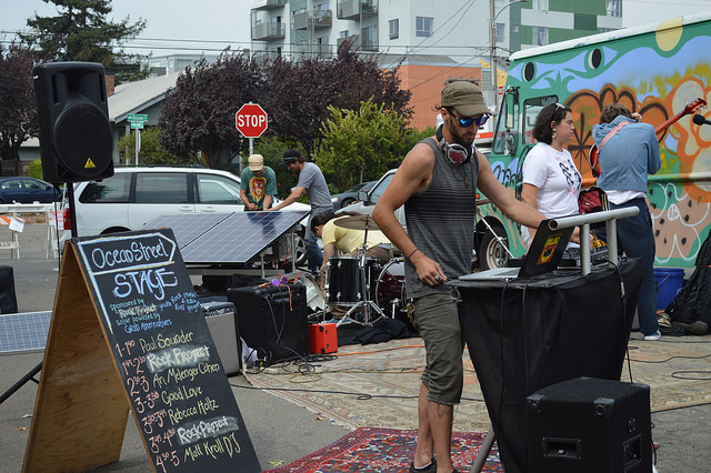 Musician performs at outdoor street event with colorful painted van and "Oceanside Skye" sign visible