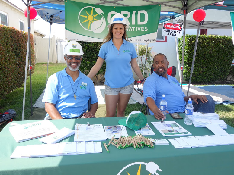 Three GRID Alternatives staff members at outdoor booth with green table, materials, and water bottles