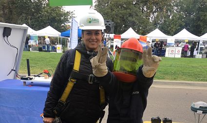 Two people in safety gear at an outdoor event, one wearing a white hard hat and yellow safety vest, the other in an orange hard hat and red vest, both giving thumbs up