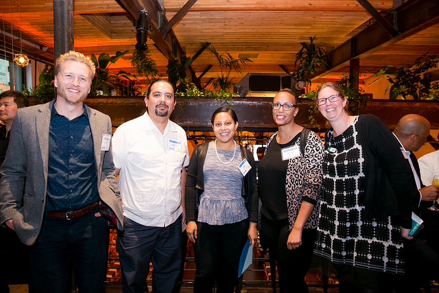 Six people standing together at an indoor event with wooden beams and plants visible in the background