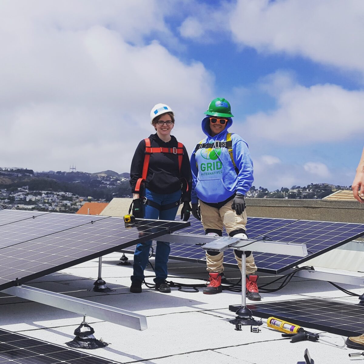 Two workers in safety gear stand on a rooftop solar panel installation overlooking a residential neighborhood