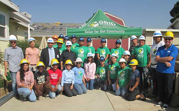 Large group of students and adults wearing hard hats and green "GRID" shirts at solar installation project under green tent banner