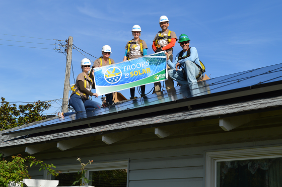 Solar workers in hard hats holding "Troops to Solar" banner on residential roof