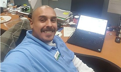 Man in blue shirt smiling at desk with laptop and workspace materials