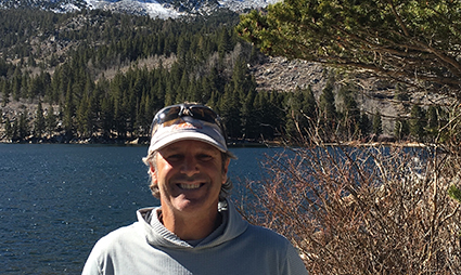 Man in white cap smiling at alpine lake with forested mountains in background