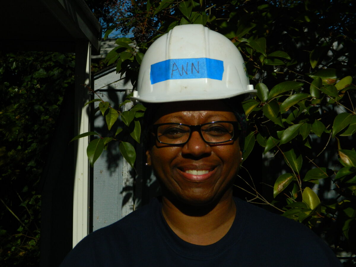 Person wearing white hard hat with "ANN" written on blue tape, smiling at camera surrounded by green foliage