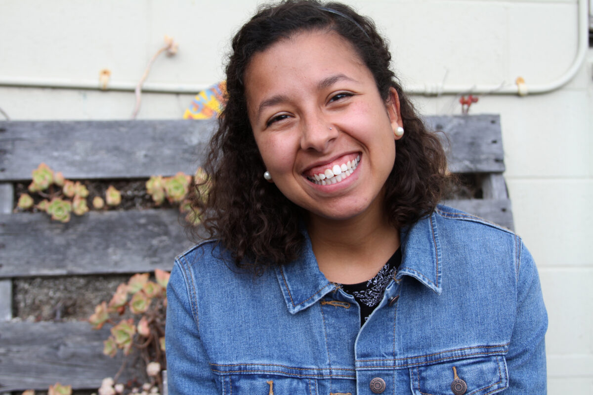 Smiling woman with curly dark hair wearing a denim jacket in front of a wooden planter box