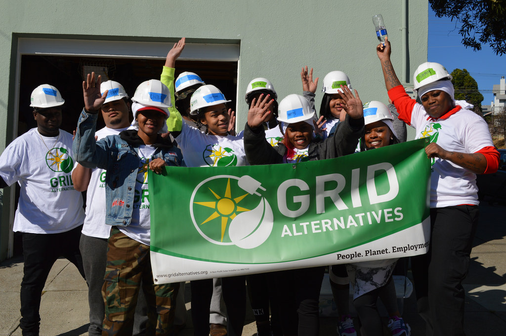 Group of workers wearing white hard hats holding a green GRID Alternatives banner outdoors