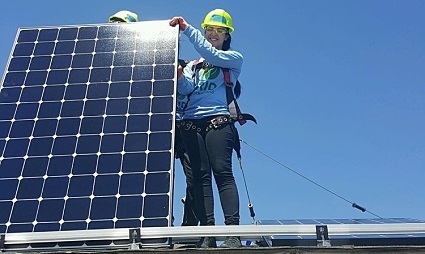 Worker in safety gear installing solar panel on rooftop against blue sky