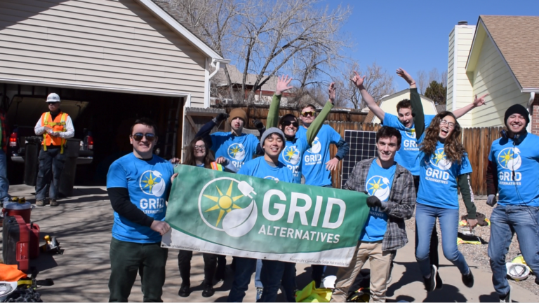 Group of volunteers in blue GRID Alternatives shirts holding a banner in front of a house during a solar installation project