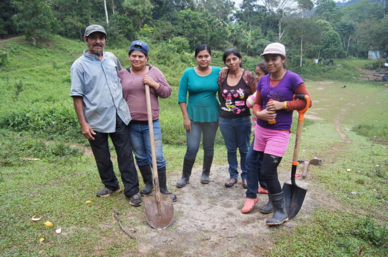 Group of five people holding shovels in a grassy field with forested hills in background