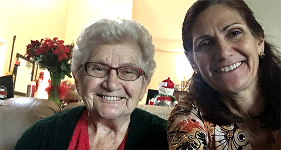 Two smiling women at a holiday gathering, older woman with white hair and glasses, younger woman with dark hair