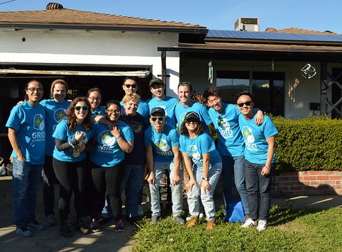 Large group of people wearing matching blue t-shirts posing outside a house on a sunny day