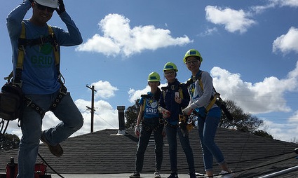 Four construction workers in hard hats stand on a residential roof during a solar installation project