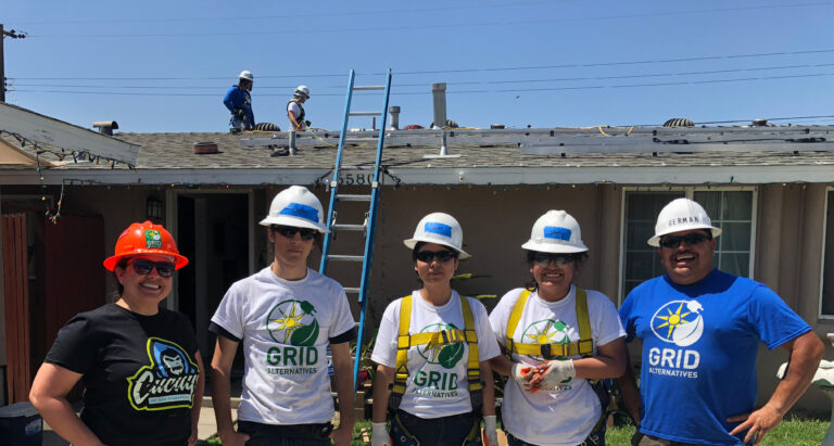 Five volunteers in safety gear and GRID Alternatives shirts stand before a house with workers on the roof