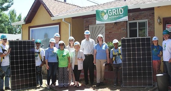 Group wearing hard hats poses with solar panels in front of house with GRID Alternatives sign