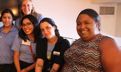 Five diverse women smiling together indoors, wearing purple and black work shirts, for GRID's "How I Got Here" series featuring Ashley Christy