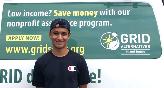 Young man in black shirt standing in front of GRID Alternatives Inland Empire nonprofit banner promoting assistance program