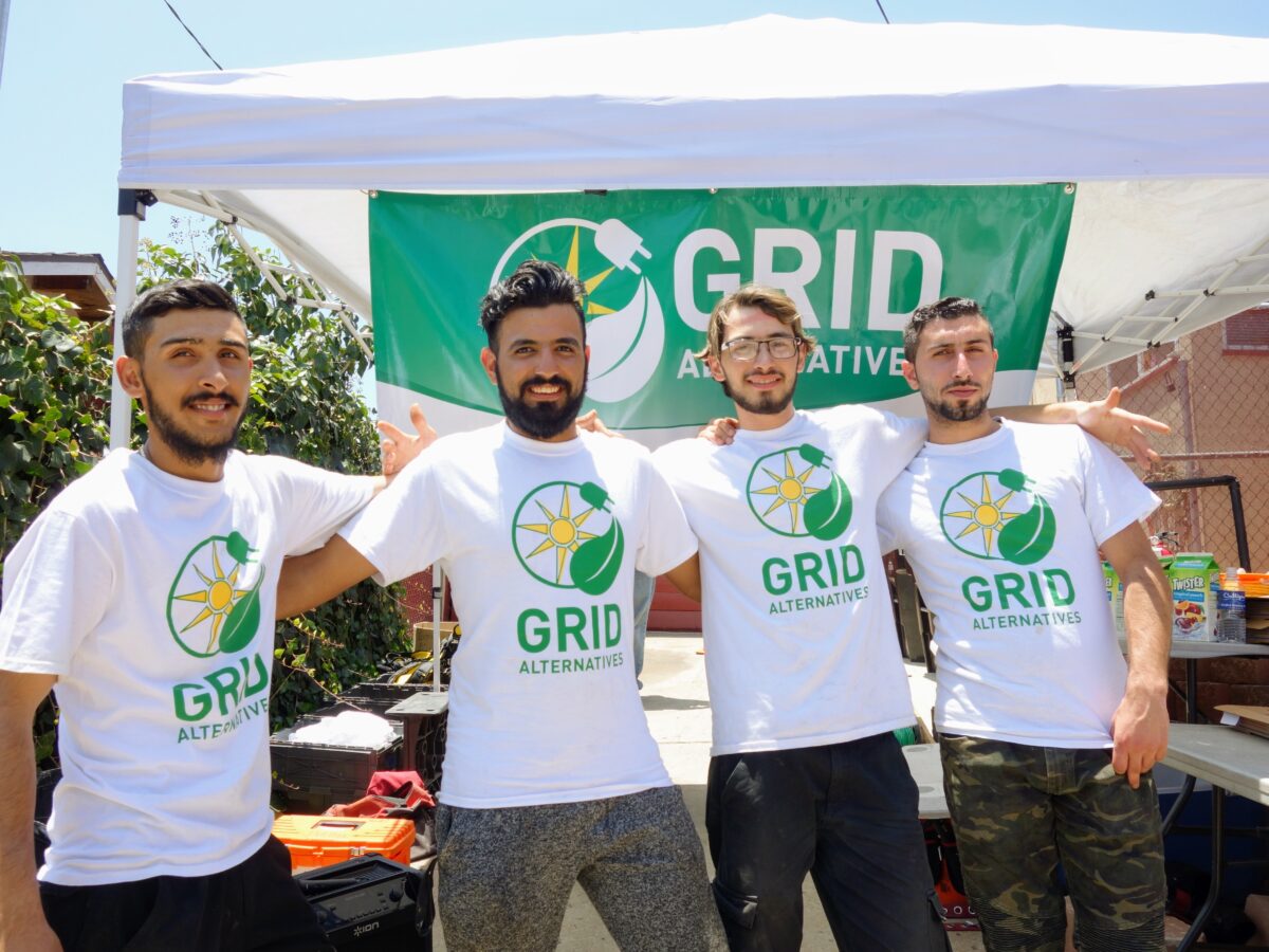 Four men wearing GRID Alternatives white t-shirts pose together under a tent at an outdoor event