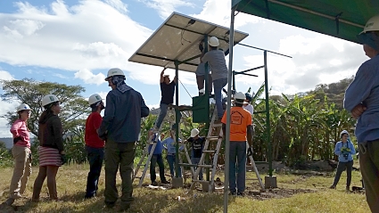 Solar panel installation demonstration with community members gathered around a structure in a rural field